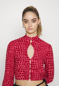 Top curto vermelho com padrão de losangos, mangas longas, decote em forma de buraco de chave e fechamento com botões na parte da frente. Feito de um material suave.
