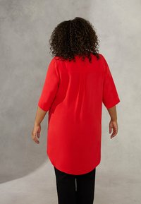 Rote, lange, leichte Bluse mit einem Rückenschlitz und Dreiviertelärmeln. Kombiniert mit schwarzen Hosen, von hinten betrachtet.