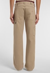Béžové cargo kalhoty s volným střihem, které mají dvě zadní kapsy, boční kapsu a viditelné švy. Materiál je odolná bavlna.