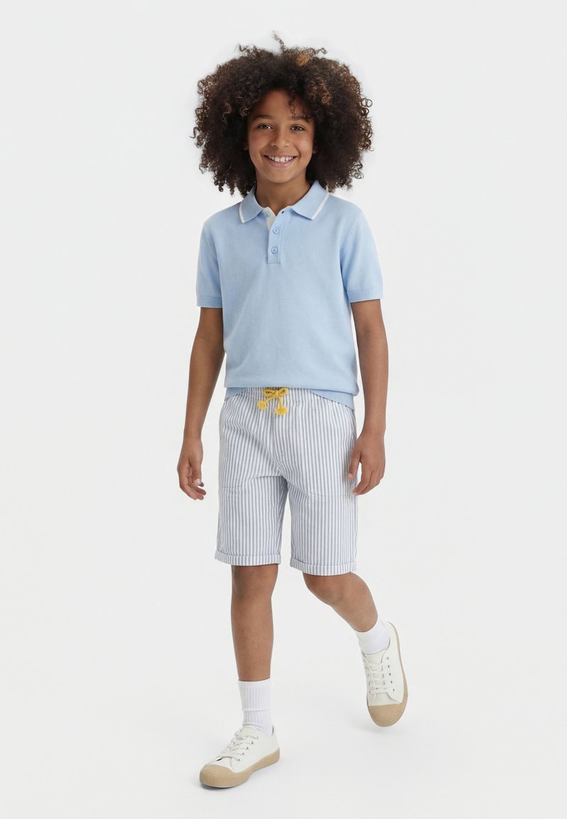 Niño sonriente con cabello rizado caminando, vestido con una camiseta tipo polo azul claro, pantalones cortos a rayas con cordón amarillo, calcetas blancas y zapatillas beige.