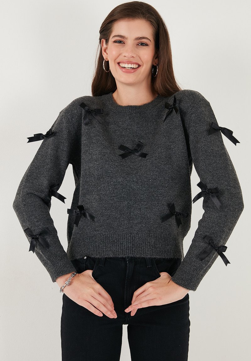 Grijze gebreide trui met zwarte satijnen strikken aan de mouwen. Geribbelde onderkant en halslijn. Gekoord met zwarte high-waisted jeans.