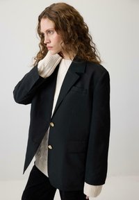 Melns žakets ar zeltītām pogām, strukturēts dizains, ar vienu krūšu kabatu, uzvilkts virs krēmkrāsas adīta džempera ar ribotiem piedurknēm.