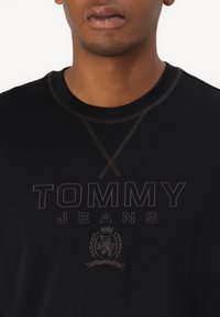 Černé bavlněné tričko s kulatým výstřihem, s logem "TOMMY JEANS" a vyšitým emblémem tmavě hnědé barvy na prsou.