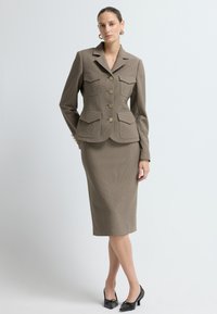 Blazer ajusté à carreaux marron avec cinq boutons et deux poches avant, associé à une jupe crayon assortie. Des chaussures à talons noires complètent le look.