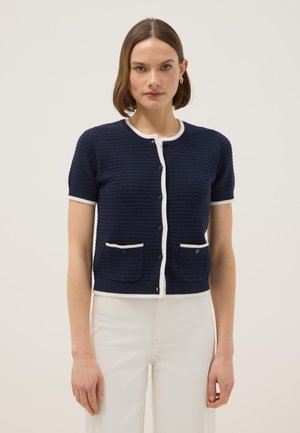 Woman wearing a navy short-sleeve buttoned cardigan with white trim and two front pockets, paired with white pants.