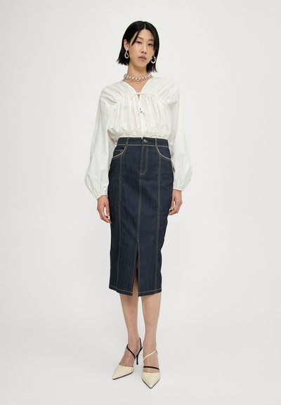 Jupe crayon en denim foncé avec une fente à l'avant, des coutures jaunes contrastantes et une taille haute. Associée à un blouse blanche à manches bouffantes et des talons.