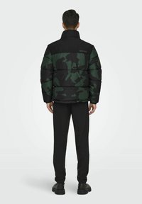 Giubbotto imbottito in camouflage nero e verde con colletto alto, dotato di sezioni trapuntate e polsini elasticizzati, abbinato a pantaloni e scarpe nere.