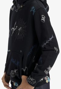 Svart hoodie med olika flerfärgad broderade mönster, inklusive tärningar, lågor, ett spindelnät och en ros. Har en framficka.