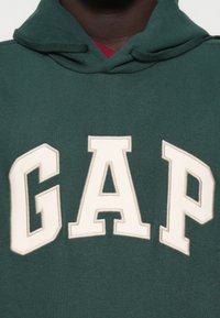 Zielona bluza z kapturem z dużym, kremowym logo "GAP". Tkanina wydaje się być miękka, z teksturowanym kapturem i warstwową czerwoną koszulą pod spodem.