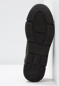 Semelle noire d'une chaussure avec des motifs de grip texturés reposant verticalement contre un mur et un sol blancs.