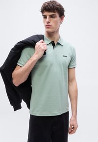 Νέος άνδρας που φοράει ανοιχτό πράσινο πόλο BOSS, κρατώντας ένα μαύρο σακάκι στον ώμο του, κοιτάζοντας προς το πλάι μπροστά σε απλό φόντο.