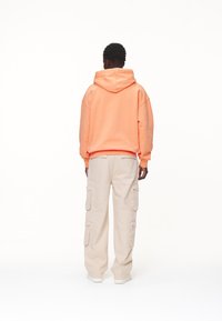 Nadrozměrná korálová mikina s kapucí na stažení, doplněná béžovými cargo kalhotami s několika kapsami, pohled zezadu.