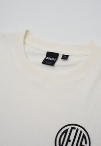 Cream-colored cotton t-shirt with a round neckline. Features a black circular logo with stylized text on the front. Tag visible at neckline.