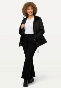 Zwarte jas met gemarmerde textuur, rits aan de voorkant en bijpassende zwarte broek. Model staat met handen in de zakken, draagt een witte top.