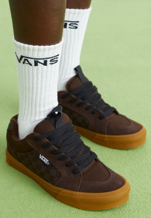 Barna velúr tornacipő fekete sakkmintával, fekete fűzővel és narancssárga gumitalppal; fehér zokni fekete "VANS" logóval.