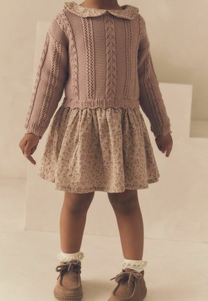 Jeune enfant portant un pull en tricot mauve, une jupe fleurie, des chaussettes à volants et des chaussures marron à lacets, debout à l'intérieur sur un fond neutre.