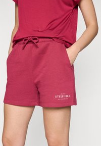 Bringebærrøde treningshorts med elastisk midje, snøring og sidelommer. Har en hvit tekstlogo nederst.