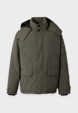 Jeune homme debout portant une doudoune vert olive sur une chemise bordeaux, un jean large bleu clair et des baskets vert olive.