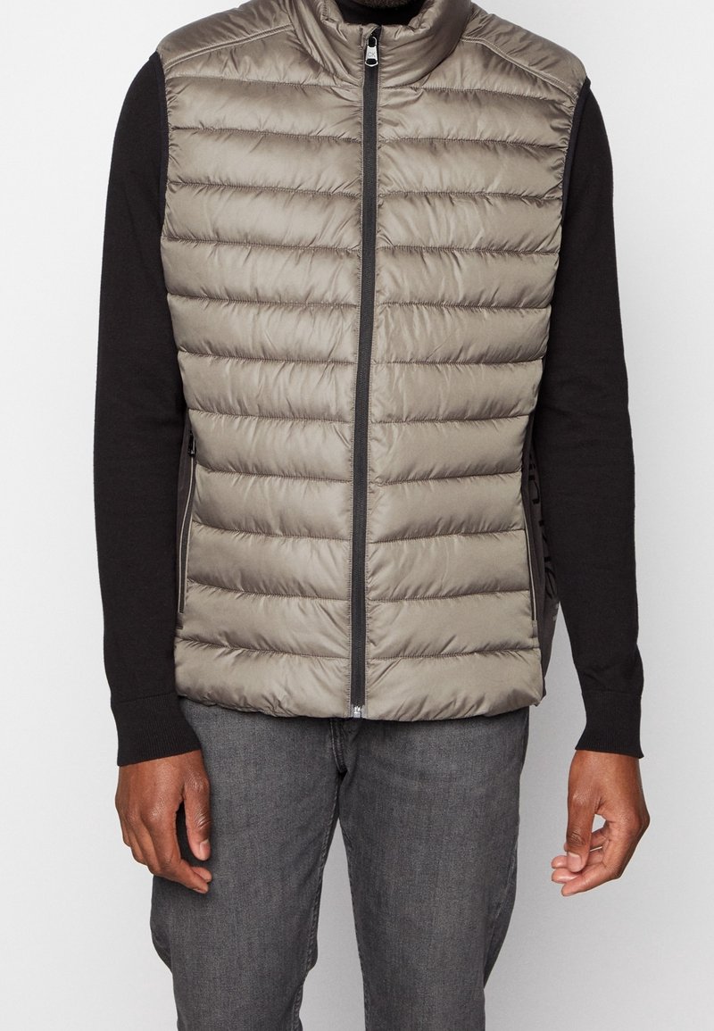Homme portant un gilet sans manches matelassé taupe avec une fermeture éclair avant, par-dessus une chemise noire à manches longues et un jean gris foncé, debout devant un fond blanc.