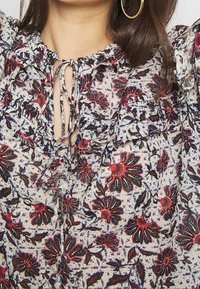Blouse à imprimé floral en tissu léger ; présente un col froncé, une fermeture à nouer, et des motifs rouges et bleus sur fond crème.