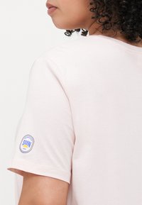 Light pink short-sleeve t-shirt made of soft cotton, featuring a circular blue and yellow logo on the left shoulder. Smooth texture, relaxed fit.