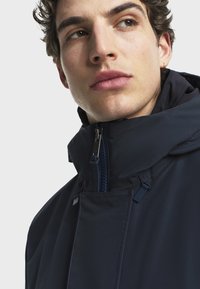 Námořnická bunda s vysokým límcem, vpředu se zipem a modrými akcenty, s hladkou texturou a minimalistickým designem.