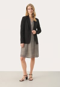 Black tailored blazer over a patterned dress with zigzag lines in black and cream. The model wears black strappy heels, standing on a neutral floor.