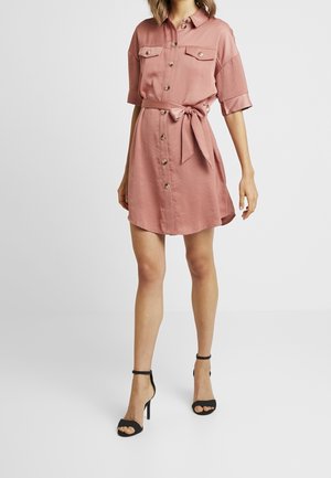 Kobieta ubrana w przybrudzoną różową sukienkę na guziki z krótkimi rękawami, wiązaną w talii, oraz czarne sandały na wysokim obcasie, stojąca na białym tle.