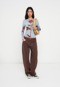 Haut bleu à manches longues avec imprimé floral rouge, associé à un pantalon large marron et des baskets noires. Porte un sac à franges beige. Fond uni.