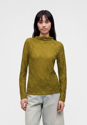 Femme aux longs cheveux foncés portant un top en dentelle vert olive à manches longues et un pantalon gris clair sur un fond clair uni.