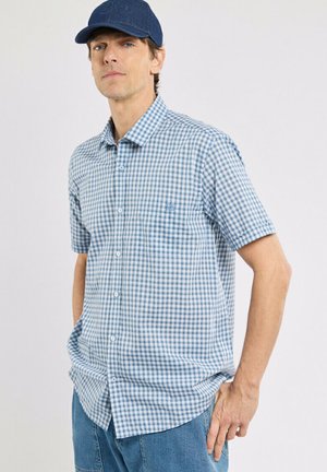 Homme portant une chemise à manches courtes à carreaux bleu et blanc, une casquette bleue et un jean bleu, debout avec une main dans la poche.