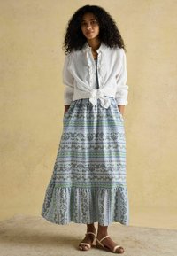 Biely ľanový top viazaný v páse cez modré maxi šaty s kvetinovým vzorom a zelenými akcentmi, s volánmi, nosené s béžovými sandálmi.