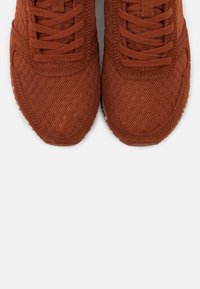 Brown mesh sneakers with suede accents, rounded toe, and flat laces. Textured surface and contrasting white sole.