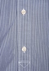 Blå och vit randig skjorta med knappslå, försedd med runda, transparanta knappar och kontrasterande sömmar längs kanterna.