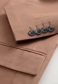 Brauner Anzugärmel mit vier braunen Knöpfen und passender Klapptasche auf hellem Hintergrund.