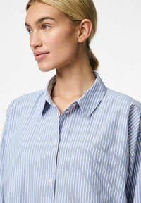 Camicia azzurra chiara con righe verticali di un blu più scuro e crema. Presenta un colletto e una tasca sul petto. Tessuto morbido al tatto.