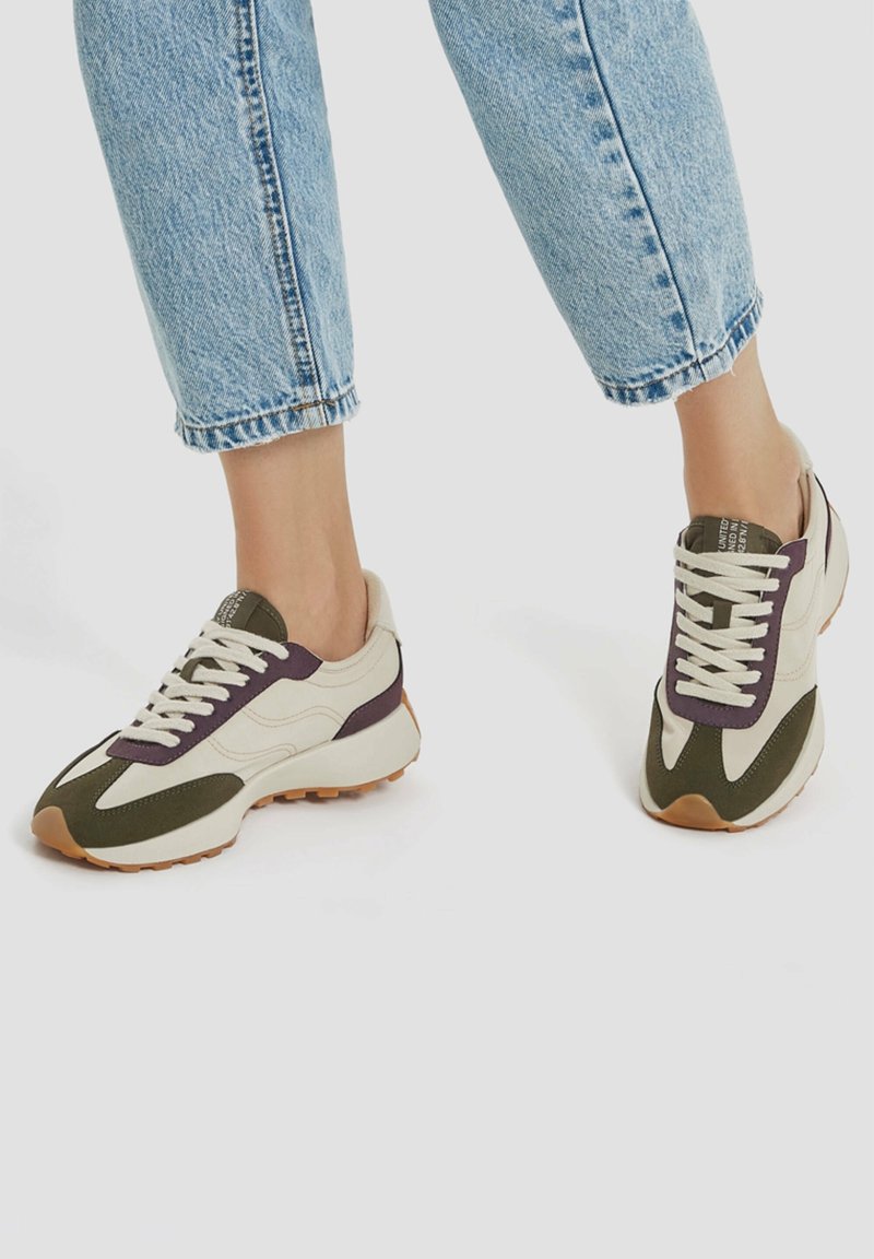 Jambes portant un jean bleu clair et des baskets crème avec des accents vert foncé et violet, debout sur un fond clair uni.