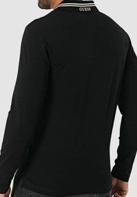 Czarna koszulka polo z długim rękawem, z białym kołnierzykiem w paski i logo „GUESS” na plecach. Wykonana z gładkiego, elastycznego materiału o dopasowanym kroju.