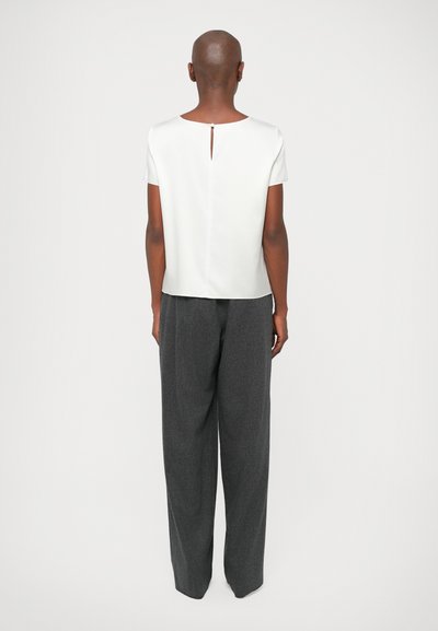 Λευκή μπλούζα κοντομάνικη με άνοιγμα στο πίσω μέρος, συνδυασμένη με φαρδιές γκρι παντελόνες, απεικονισμένη από πίσω σε φωτεινό φόντο.