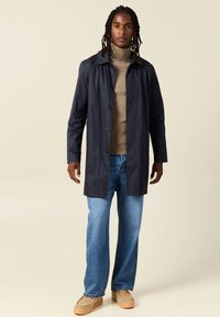 Capa impermeable en color negro marino con cuello y botones frontales, llevada sobre un suéter de cuello alto beige, combinada con vaqueros azules y zapatos beige.