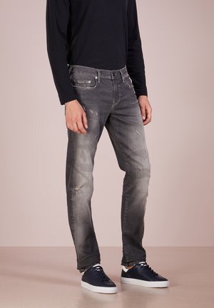 Muž ve vybledlých šedých opraných džínách, černém tričku s dlouhým rukávem a námořnicky modrých teniskách s bílými podrážkami stojí na béžové podlaze.