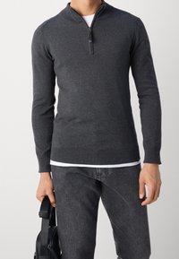 Maglione grigio lavorato a maglia con zip a quarto, polsini e orlo a coste, abbinato a jeans scuri e una borsa nera. Texture morbida e design aderente.