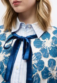 Κοντινό πλάνο γυναίκας που φοράει λευκό πουκάμισο με ανοιχτό μπλε ρίγες, μπλε σατέν παπιγιόν και υφασμάτινη ζακέτα με μεγάλα μπλε λουλουδάτα σχέδια.