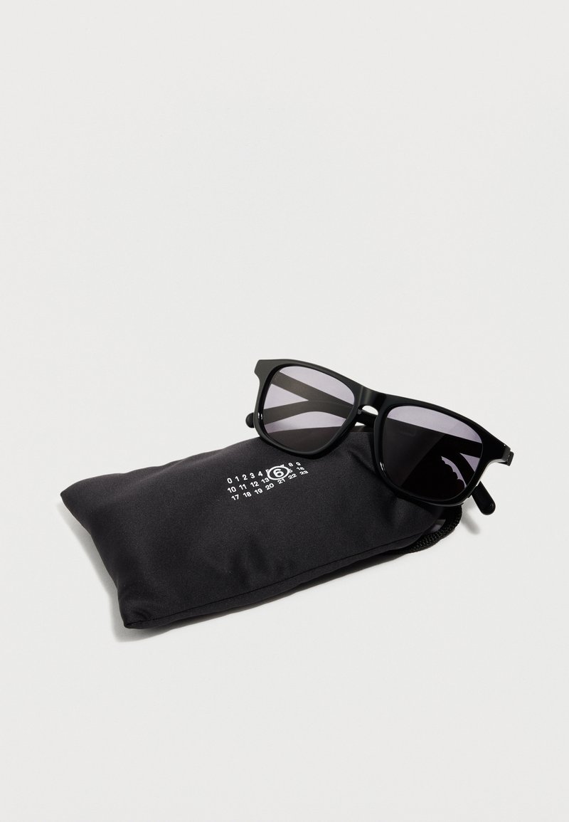 Schwarze Sonnenbrille mit dunklen Gläsern, die auf einem schwarzen, weichen Etui liegt, das ein weißes, kreisförmiges Logo mit Zahlen auf weißem Hintergrund zeigt.