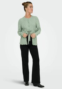 Mint zelena pletena jopica z vzorcem teksturiranih diamantov, okrogel ovratnik, zapiranje z gumbi in kais na pasu; v kombinaciji z črnimi hlačami in čevlji.