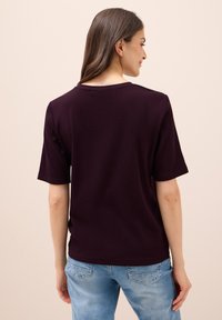 Tmavě fialový top s krátkým rukávem a kulatým výstřihem, texturovaná látka, volný střih, v kombinaci s světle modrými džíny.