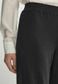 Svarta, vida byxor i mjukt stickat material, med en elastisk midja och en slät yta. En hand syns nära midjan.