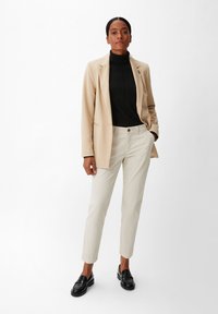 Beige Blazer über einem schwarzen Rollkragenpullover, kombiniert mit hellbeigen Hosen und schwarzen Loafers. Glatte Stoffe mit maßgeschneidertem Schnitt und minimalen Details.
