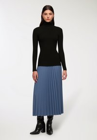 Czarny prążkowany sweter z wysokim kołnierzem w połączeniu z jasnoniebieską plisowaną maxi spódnicą. Modelka nosi czarne buty na wysokim obcasie. Gładkie tekstury materiałów.