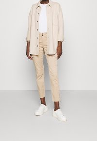 Beige košilová košile, bílý tričko, světle béžové cargo kalhoty s bočními kapsami a bílé tenisky, stojící na jednobarevném pozadí.